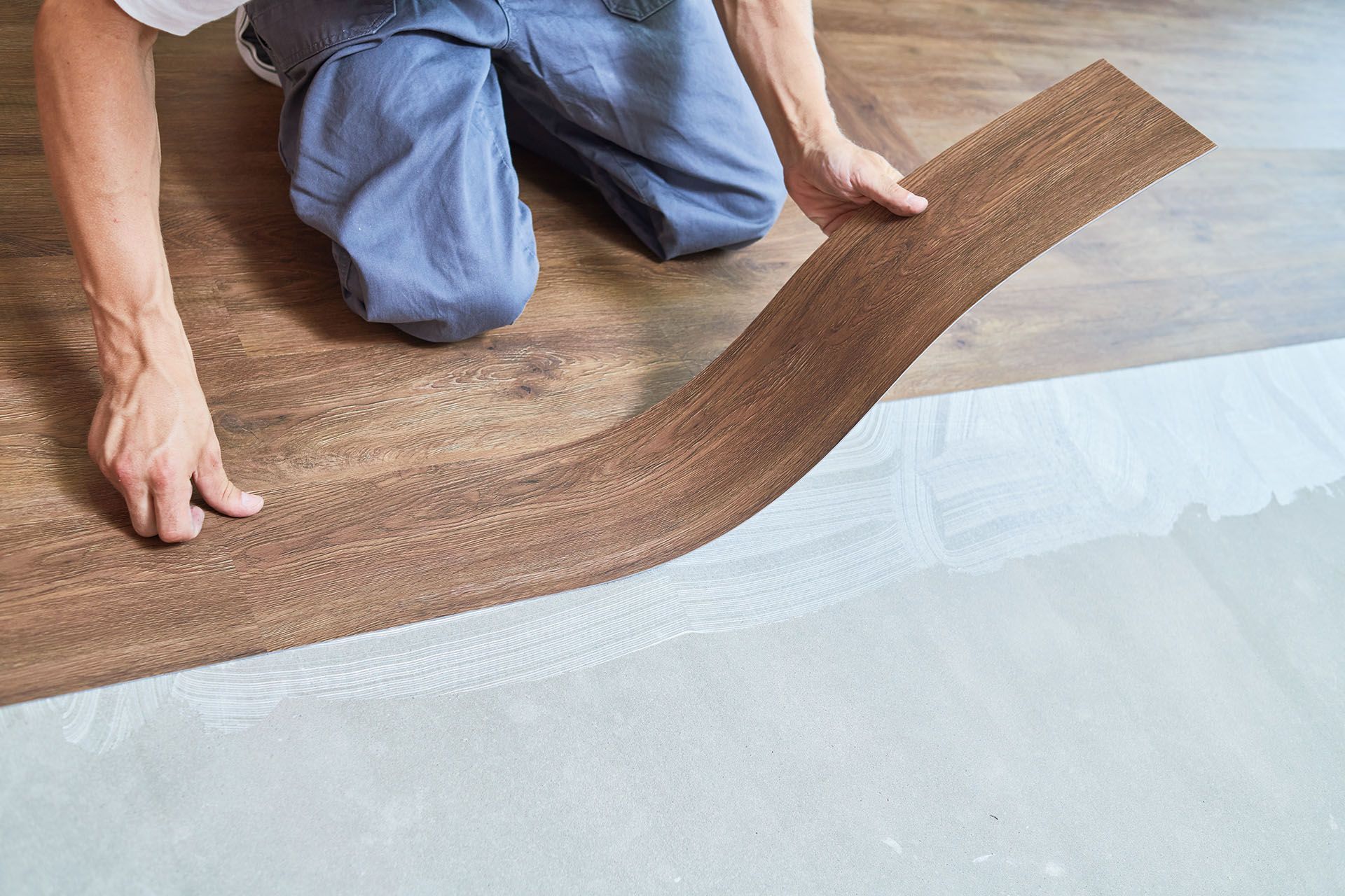 A worker installing wood-look vinyl flooring over adhesive on a concrete subfloor.