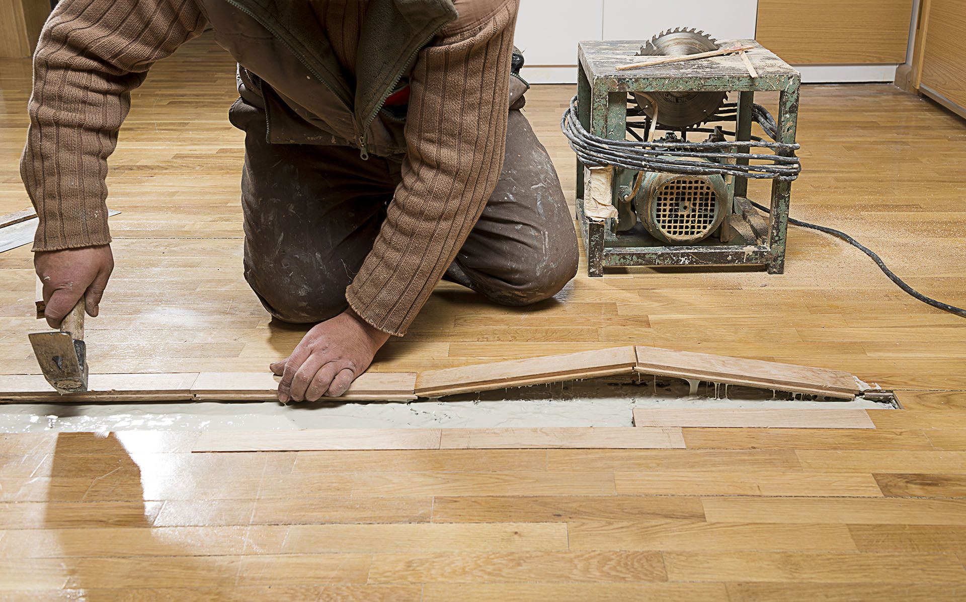 A person kneeling on a wooden floor, using a tool to repair buckled floorboards next to a table saw.