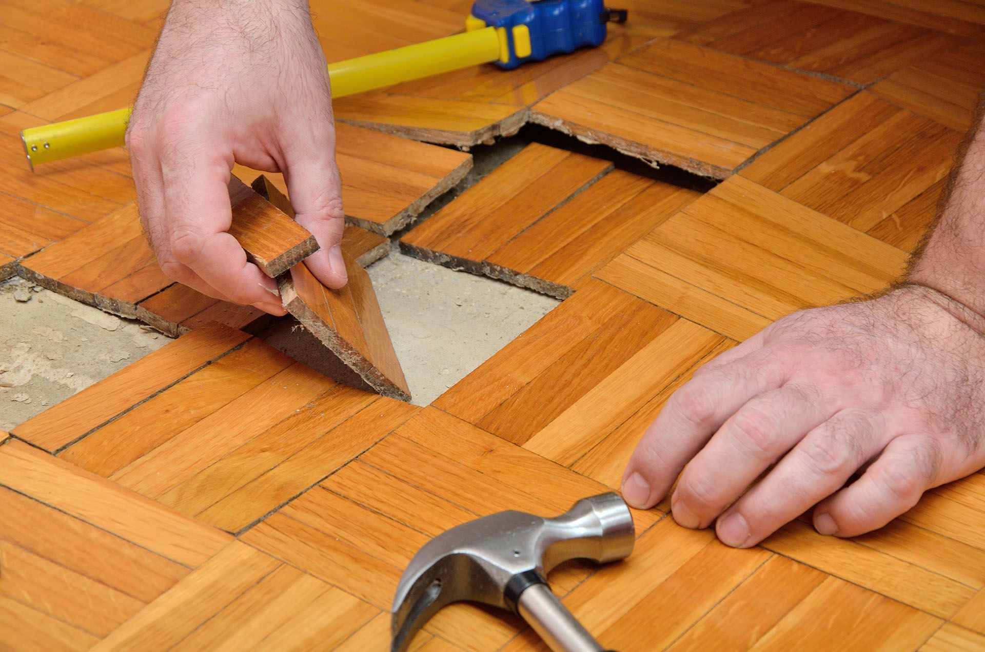 Hands repair damaged parquet wood flooring using a hammer and measuring tape.
