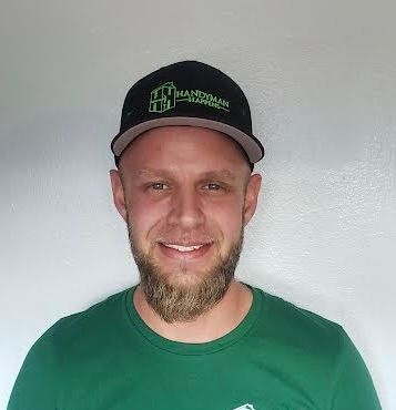 A man with a short beard and a green shirt, wearing a black baseball cap with a green logo, smiles against a gray wall.