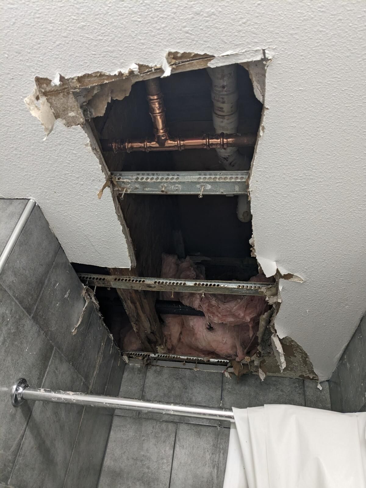 A large, irregular hole in a bathroom ceiling reveals exposed copper pipes, metal framing, and pink fiberglass insulation.