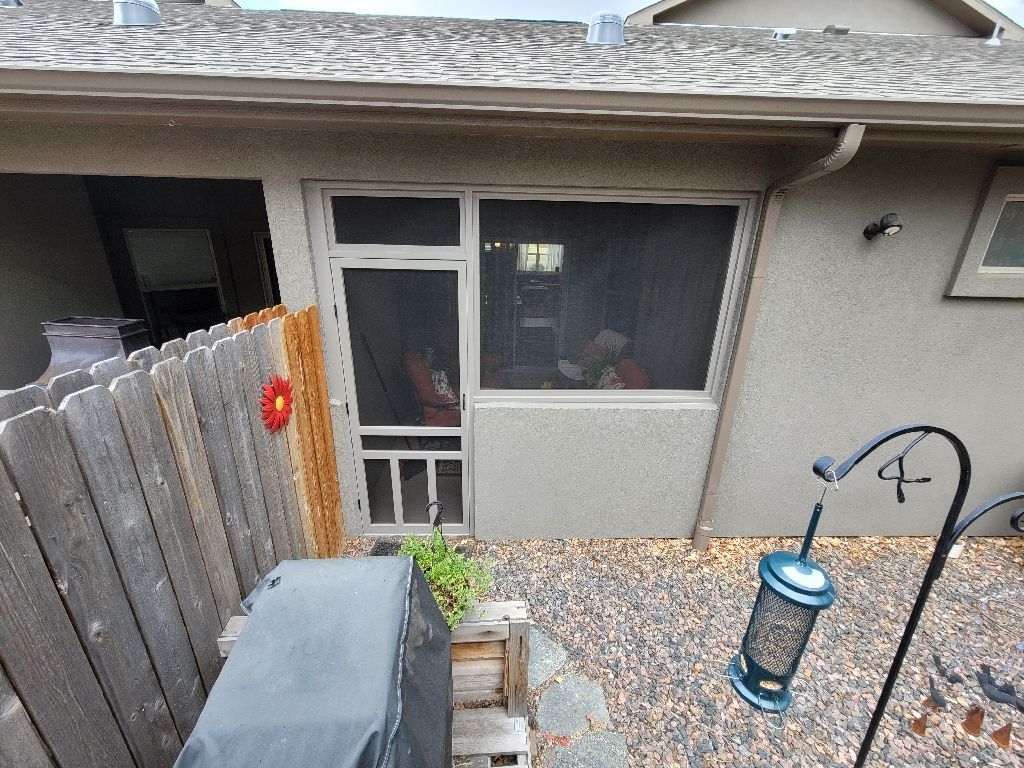 A patio area featuring a screened door and window, a wooden fence, a covered grill, and a hanging bird feeder.