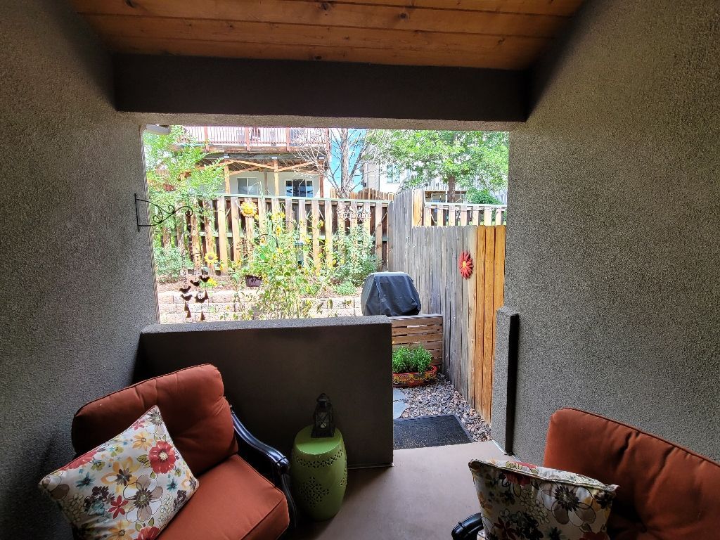 A shaded patio with two orange chairs and a small green stool overlooking a backyard with a wooden fence and greenery.
