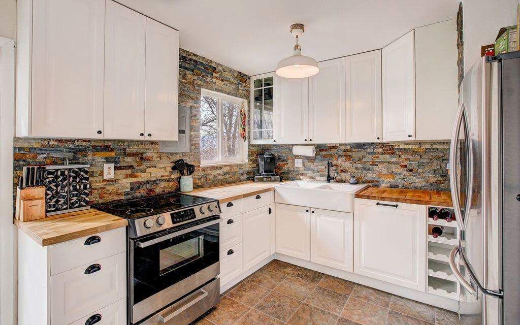Modern kitchen with white cabinets, stone backsplash, wood countertops, stainless steel appliances, and tile flooring.