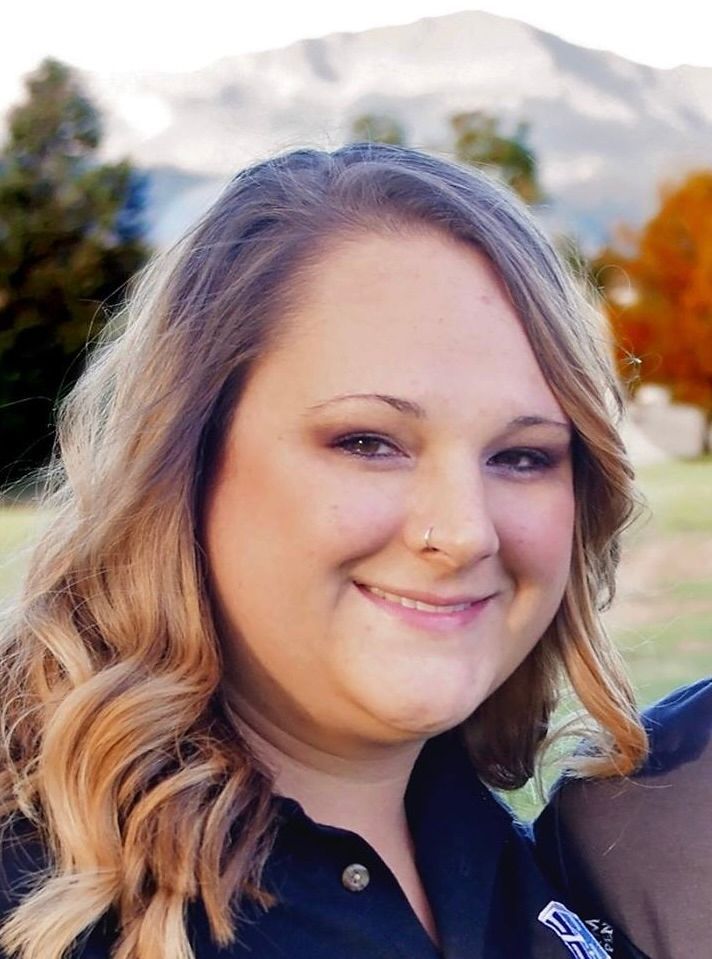 A smiling person with wavy, light brown hair wearing a dark collared shirt, outdoors with blurred trees in the background.