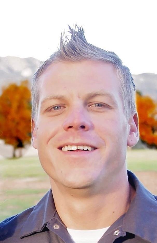 A smiling person with a spiked hairstyle, wearing a dark shirt, standing outdoors with fall foliage and mountains behind.
