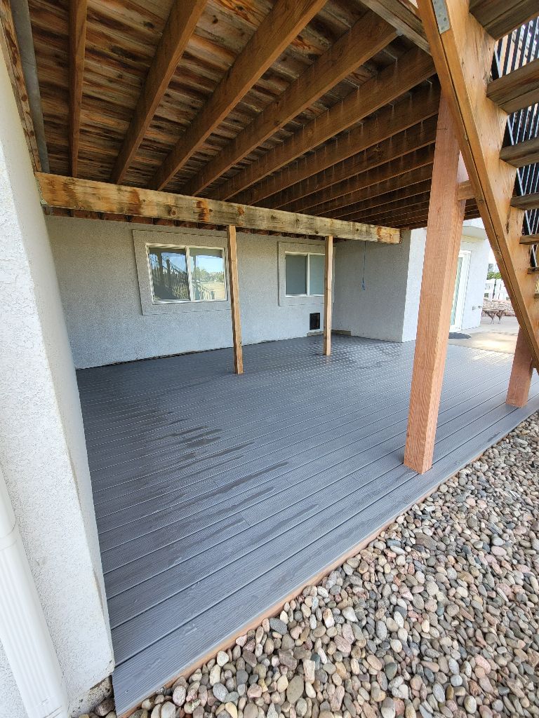 A ground-level view of a patio with grey composite decking under a wooden deck structure, next to a gravel-covered yard.