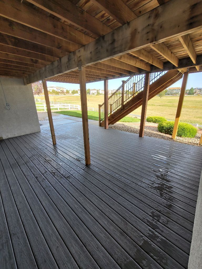 A covered outdoor patio with dark gray composite decking beneath a wooden deck structure and staircase.