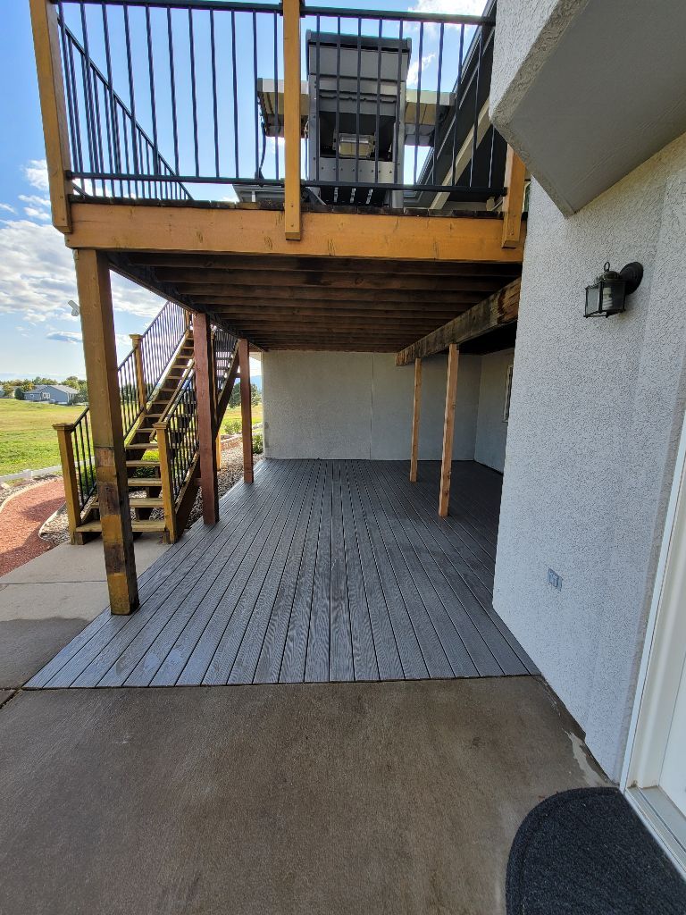 A deck over a patio with grey modular flooring, wooden stairs, and a house exterior with a wall-mounted light.