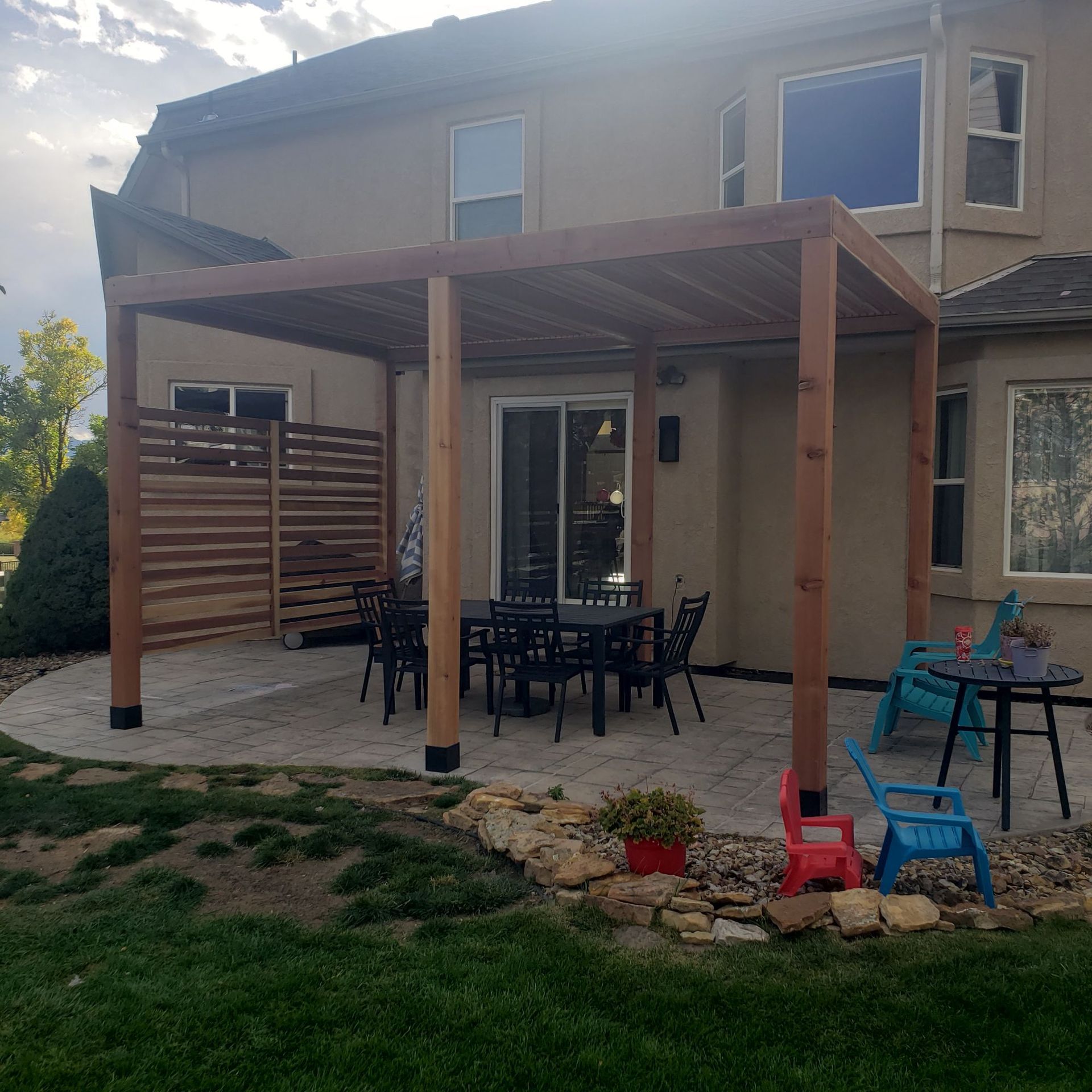 A wooden pergola covers a patio area with a dining table and chairs, adjacent to a house with a stone garden bed.