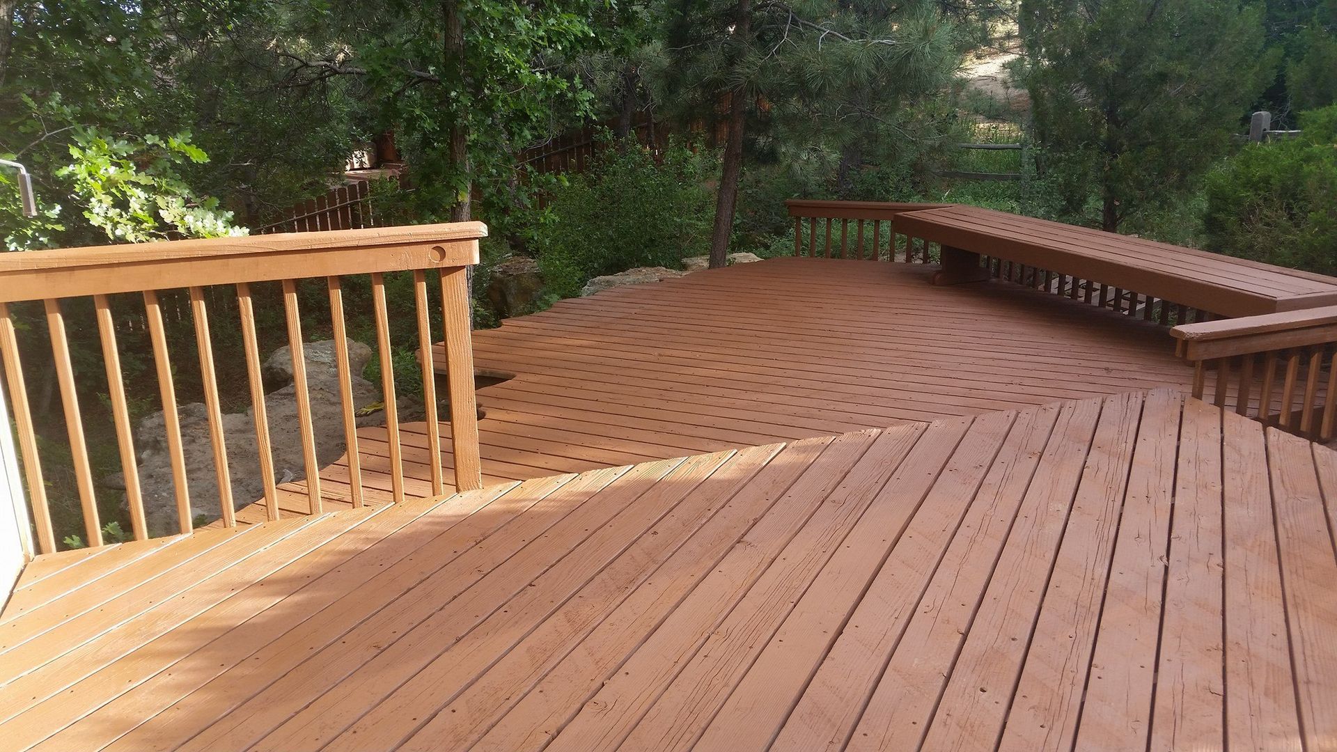 A wooden deck with straight planks and a curved, fan-patterned section, surrounded by railings and lush green trees.