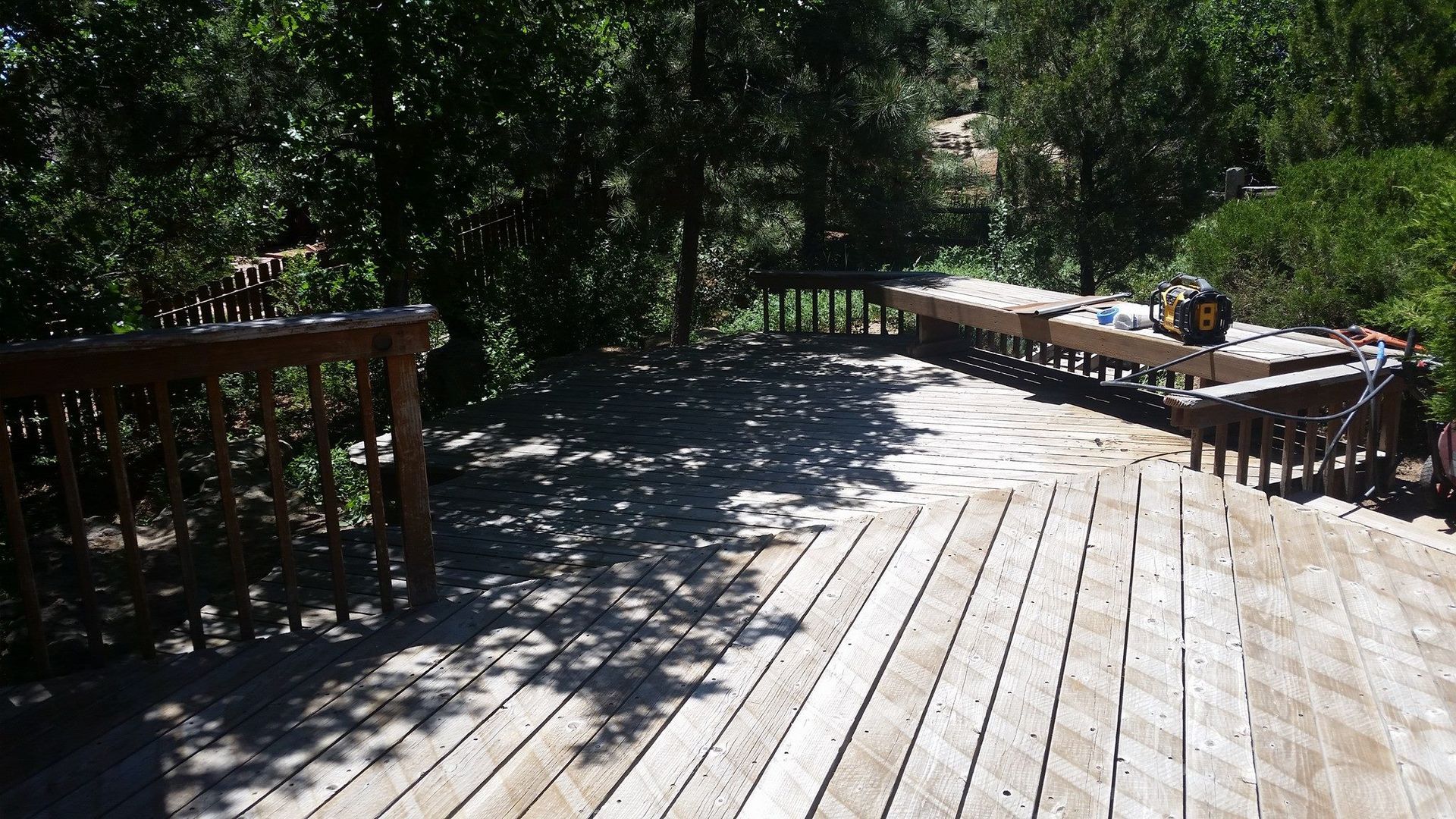 A wooden deck with railings overlooks a lush, shaded forest area on a sunny day.