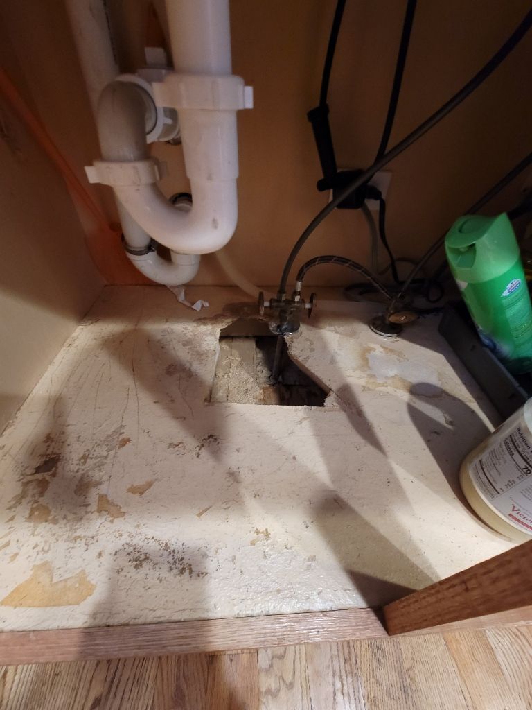 A view under a kitchen sink showing white PVC plumbing, a water shut-off valve, and a hole cut into the baseboard cabinet.