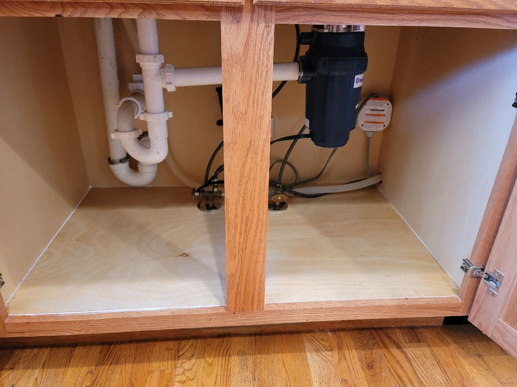 The interior of a wooden kitchen cabinet under a sink showing PVC plumbing pipes connected to a garbage disposal unit.