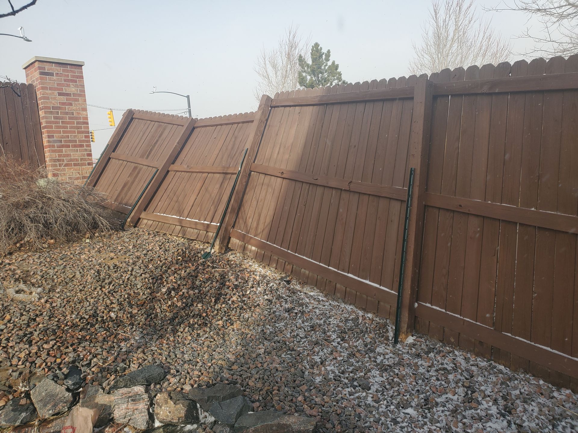 A brown wooden fence is leaning severely, tilted against a brick pillar in an outdoor yard with rocks and dry brush.