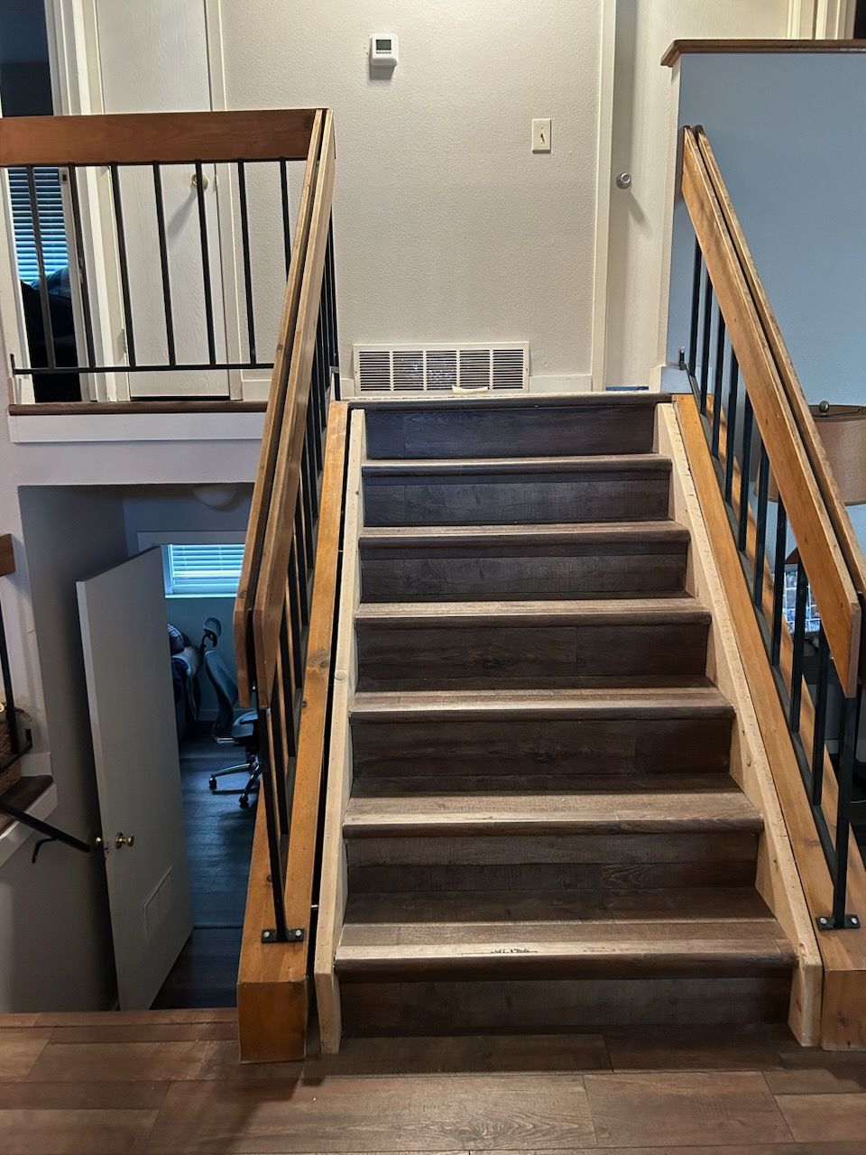 Staircase with wooden handrails and black metal spindles ascending toward a landing with a vent and thermostat.