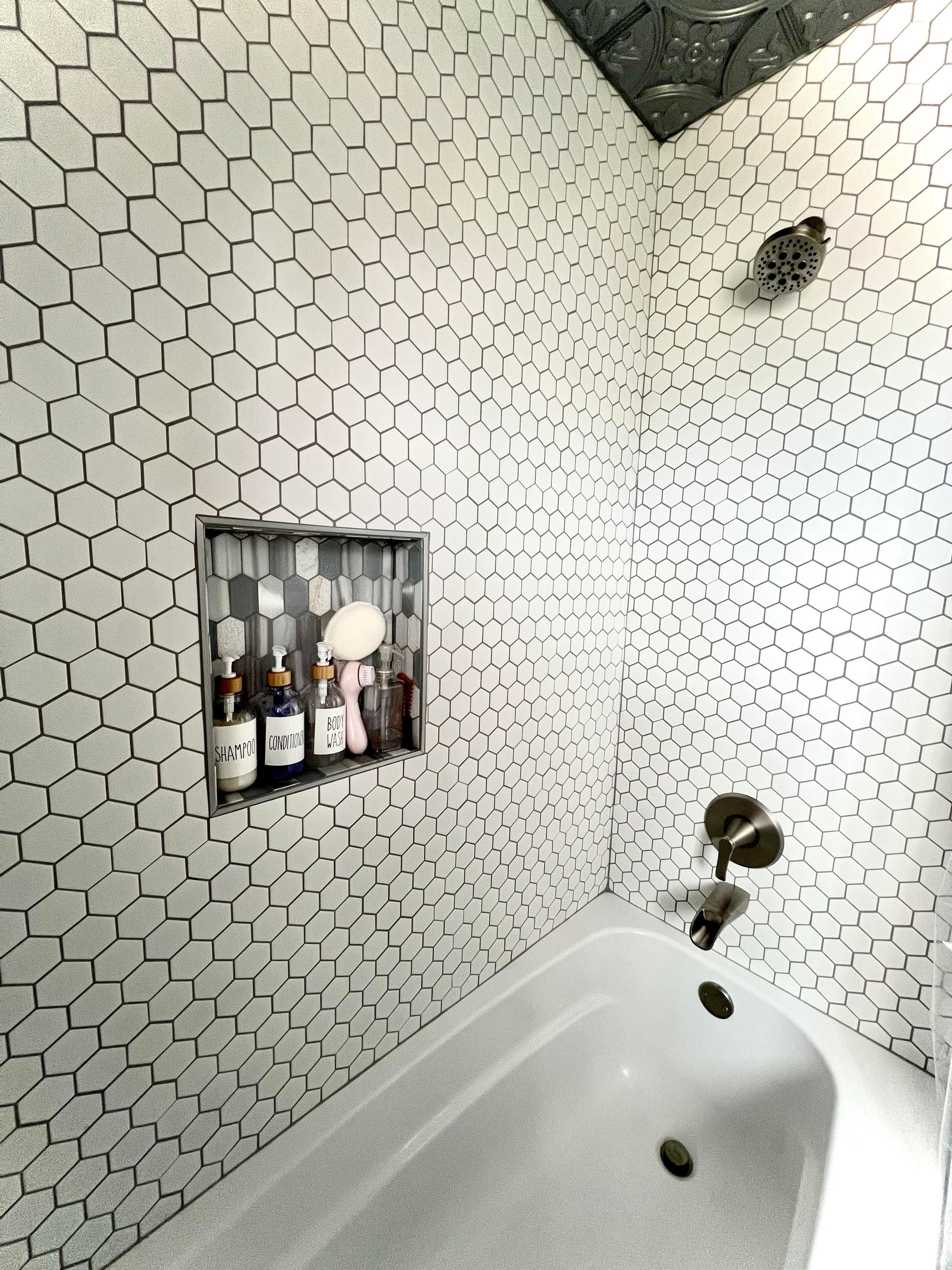 A bathtub with white hexagonal wall tiles featuring dark grout, a recessed wall niche with toiletries, and a shower head.