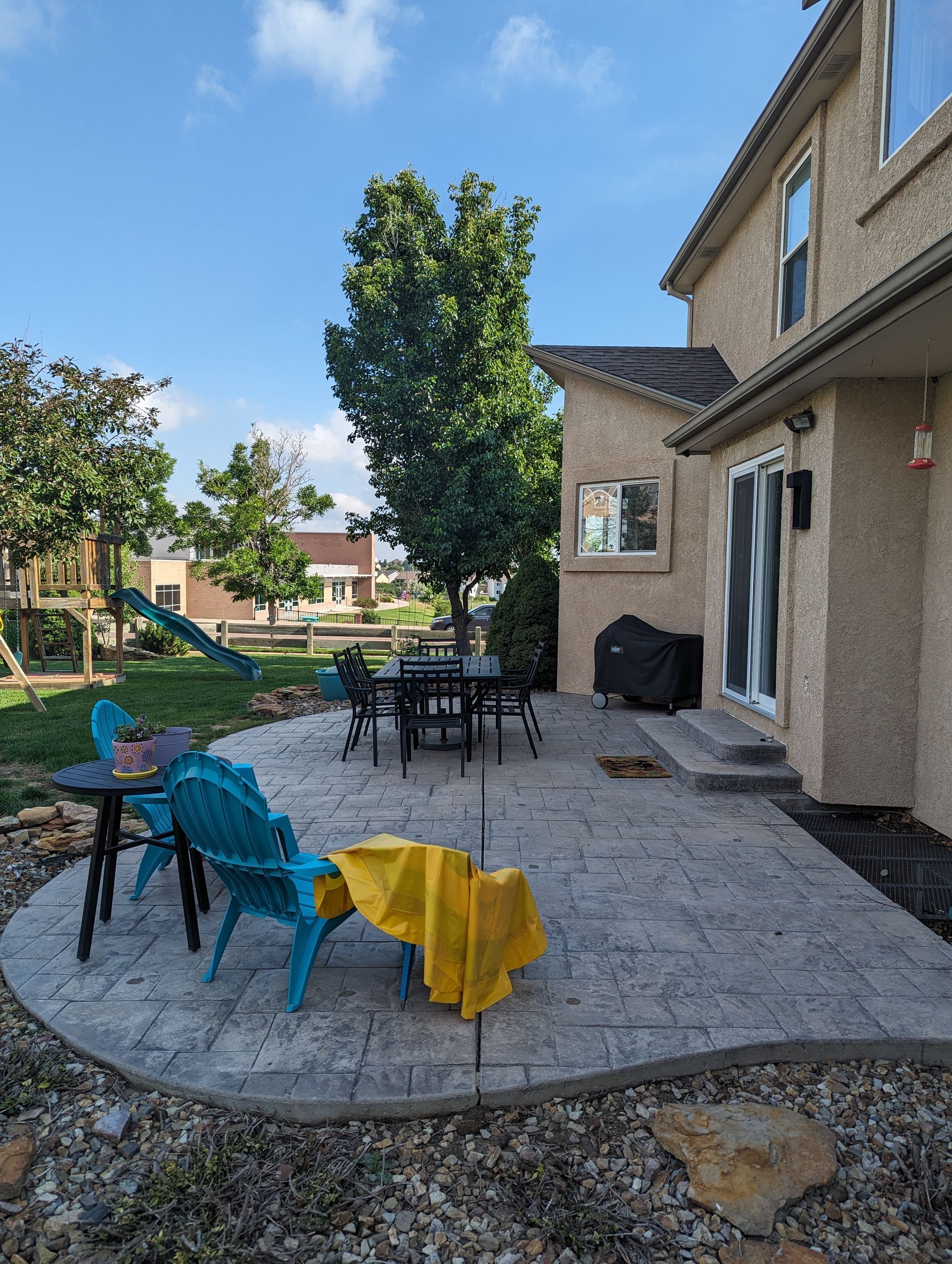 A stamped concrete patio in a backyard with a dining set, blue chairs, a grill, and a children's slide on the lawn.