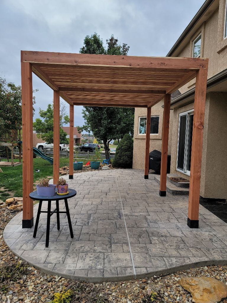 A wooden pergola sits over a stamped concrete patio next to a house, with a small round table in the foreground.