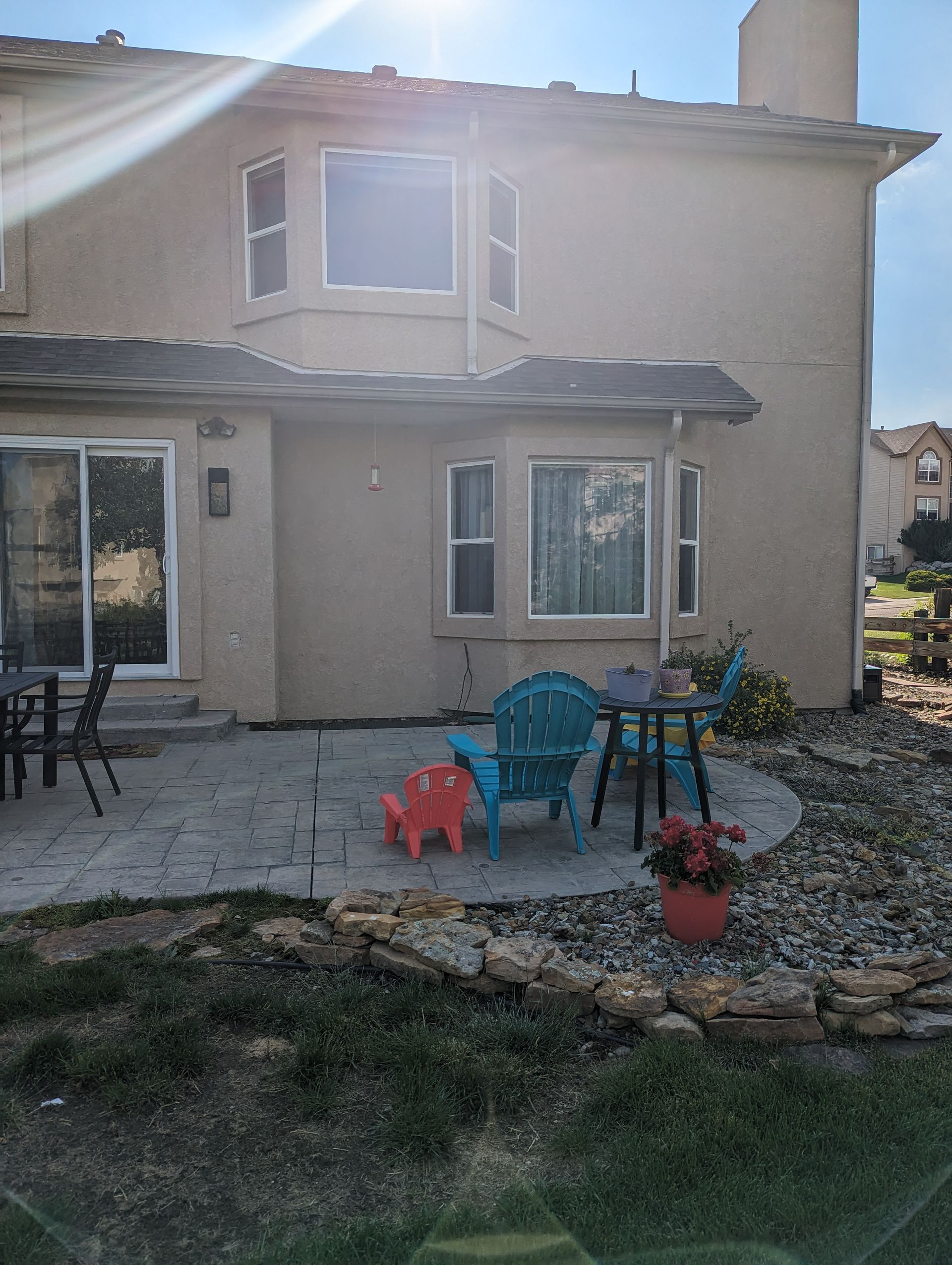 A tan house with a stone patio, featuring a teal Adirondack chair, a small red chair, and a table outdoors in the sunlight.