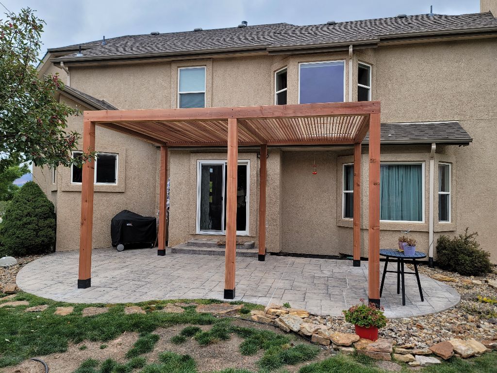 A wooden pergola stands on a stamped concrete patio in the backyard of a tan-colored, two-story house.