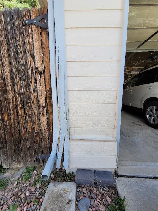 A damaged light blue downspout on the side of a beige garage, next to a wooden fence and a driveway.
