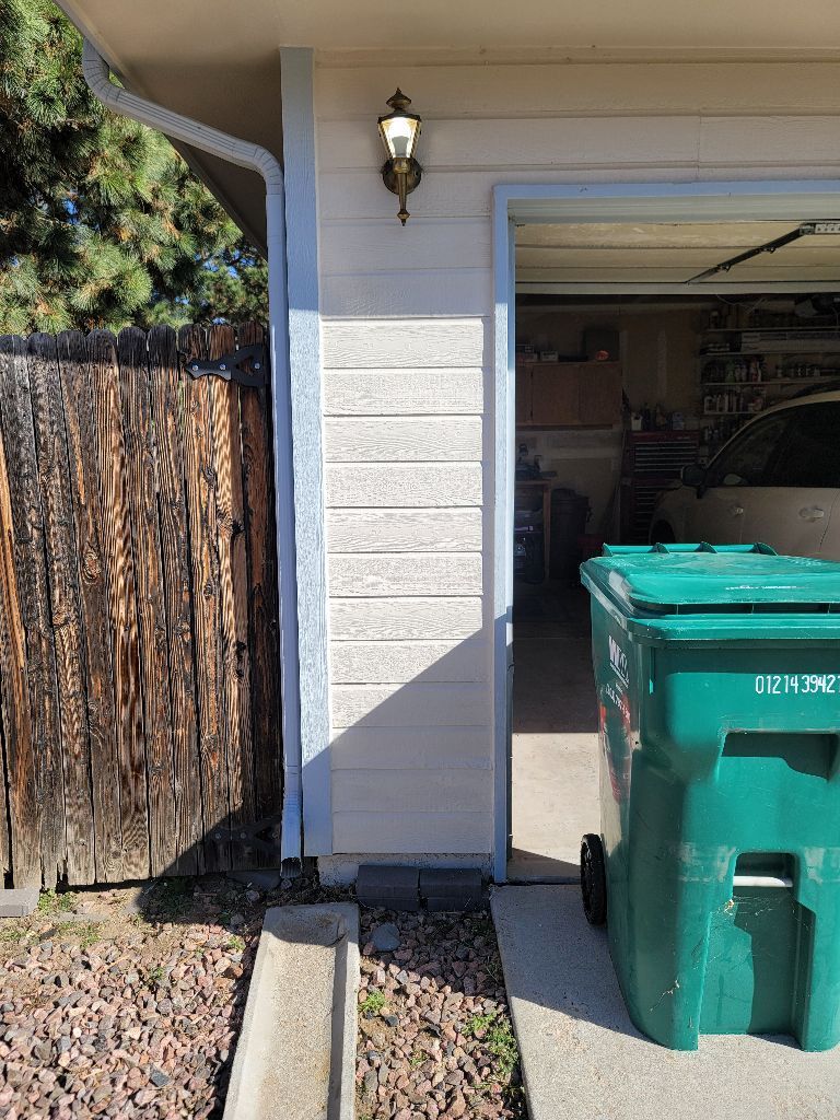 Exterior view of a beige garage wall with a light fixture, a wood fence on the left, and a large green trash bin.