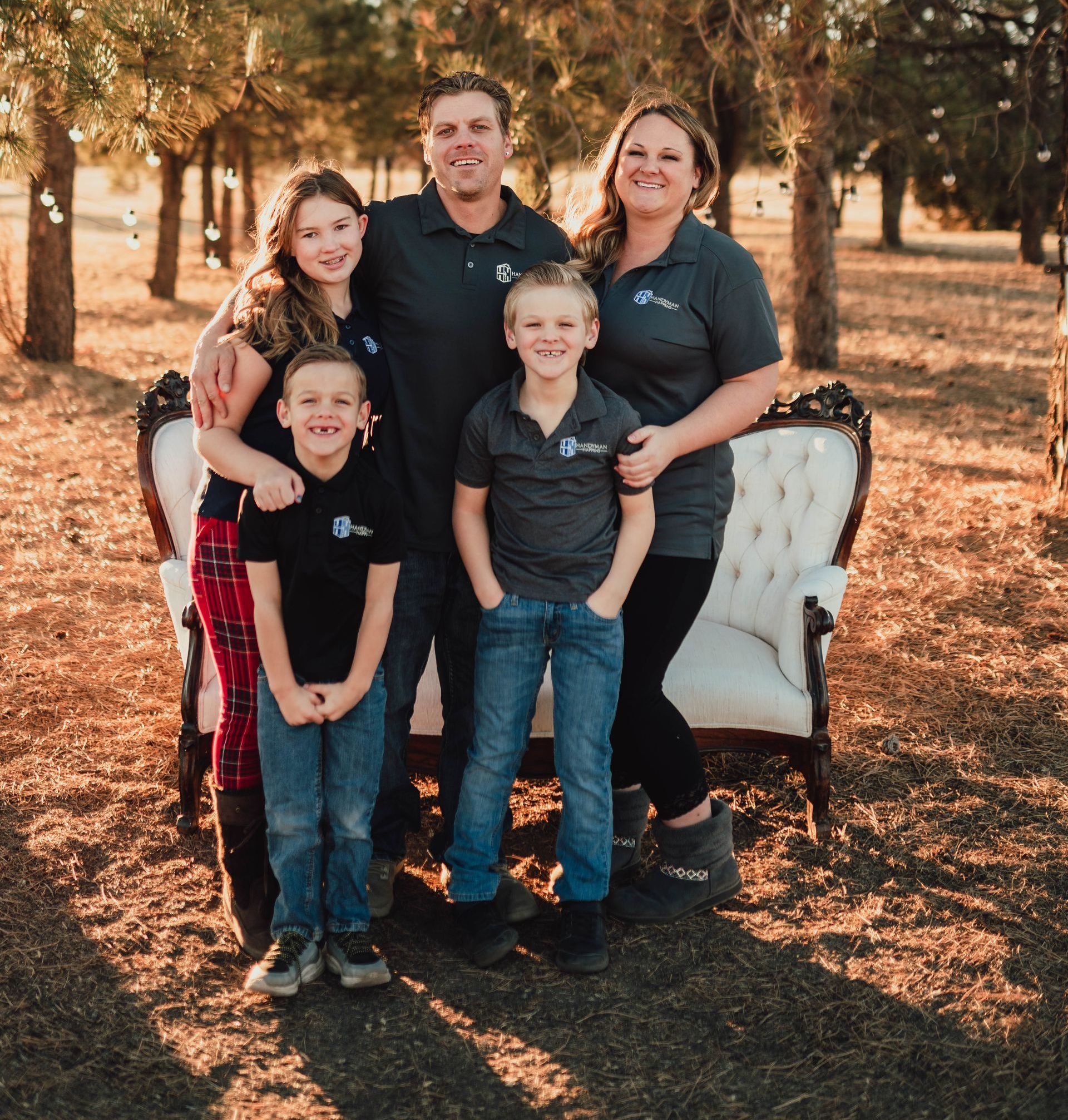 A family of five poses together outdoors in front of a white tufted bench in a wooded area during golden hour.
