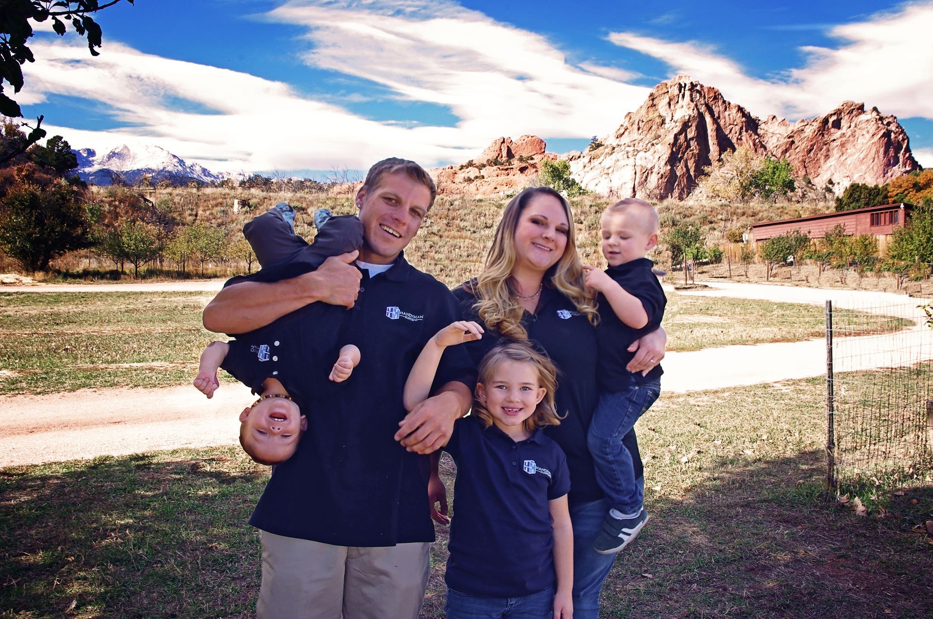 A family of five posing outdoors with mountains in the background, one child held upside down, all smiling at the camera.