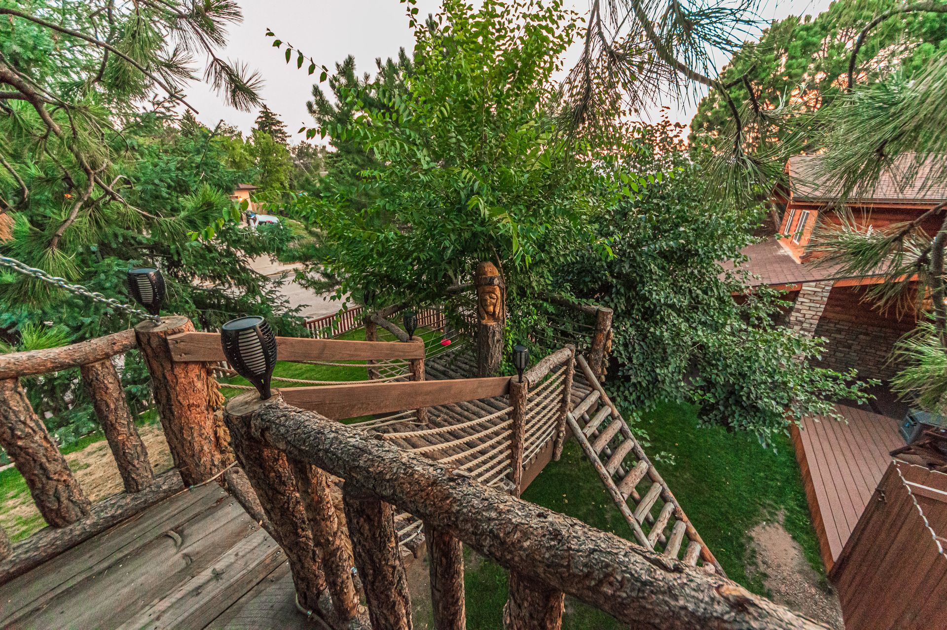 A wooden bridge with rope railings connecting two structures in a lush, green, tree-filled outdoor setting.