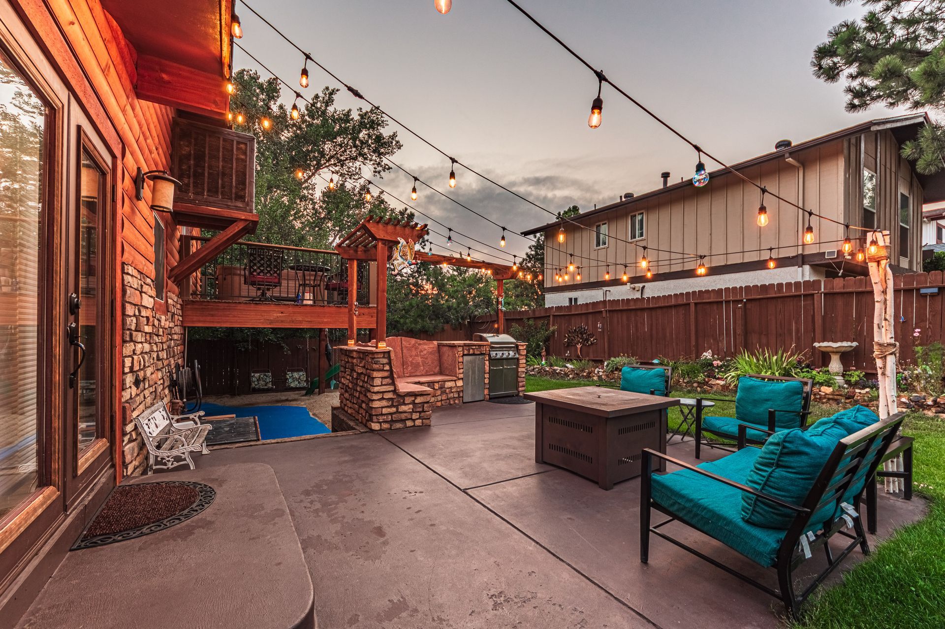 A backyard patio at dusk with string lights, a fire pit, turquoise outdoor furniture, and a stone outdoor kitchen area.