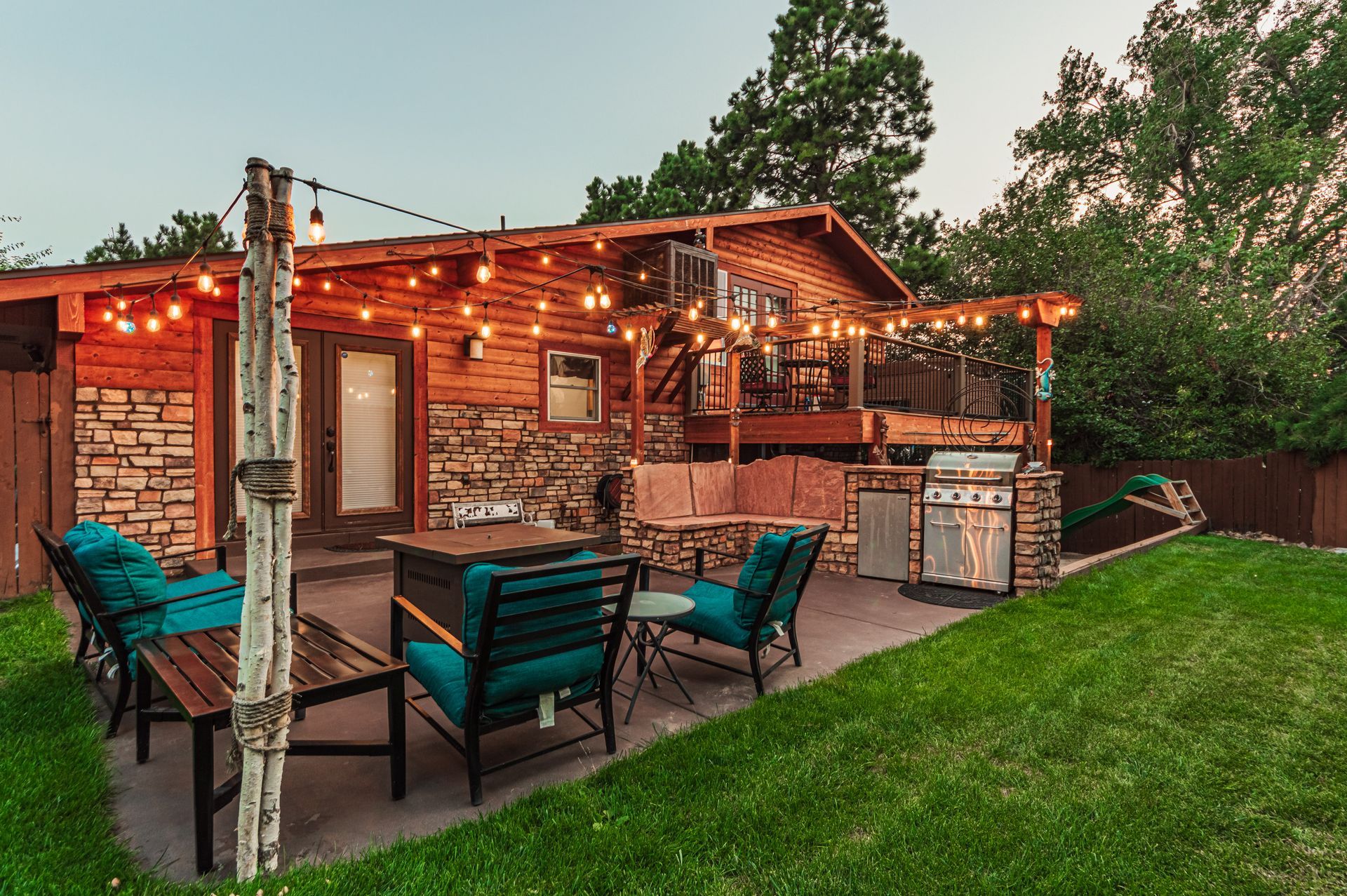 A cozy wooden cabin patio at dusk, featuring teal chairs, a fire pit, string lights, and a stone-built grill.