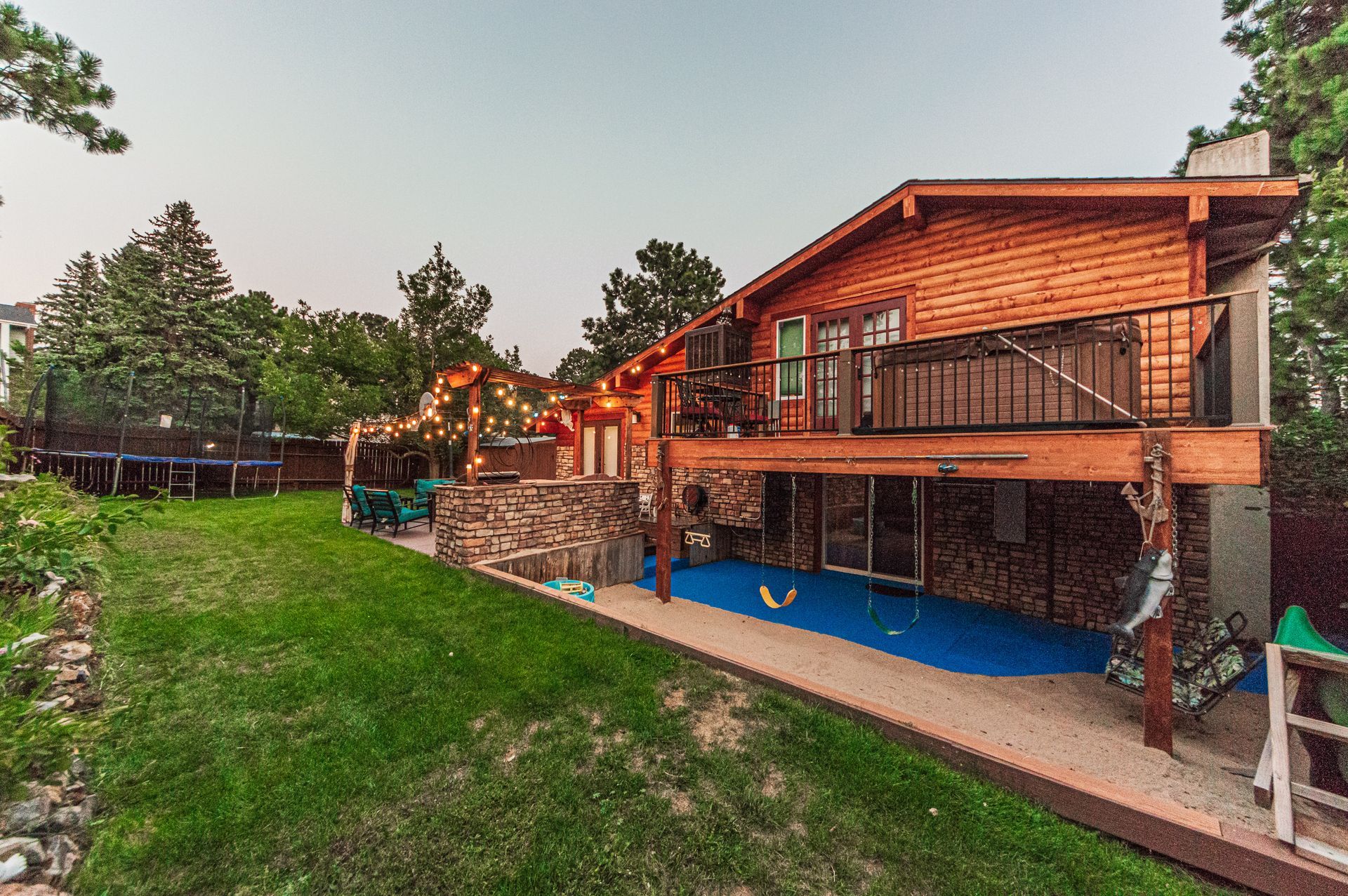 A wood-sided cabin with a deck, a hot tub, and a blue patio area overlooking a green yard with a trampoline.