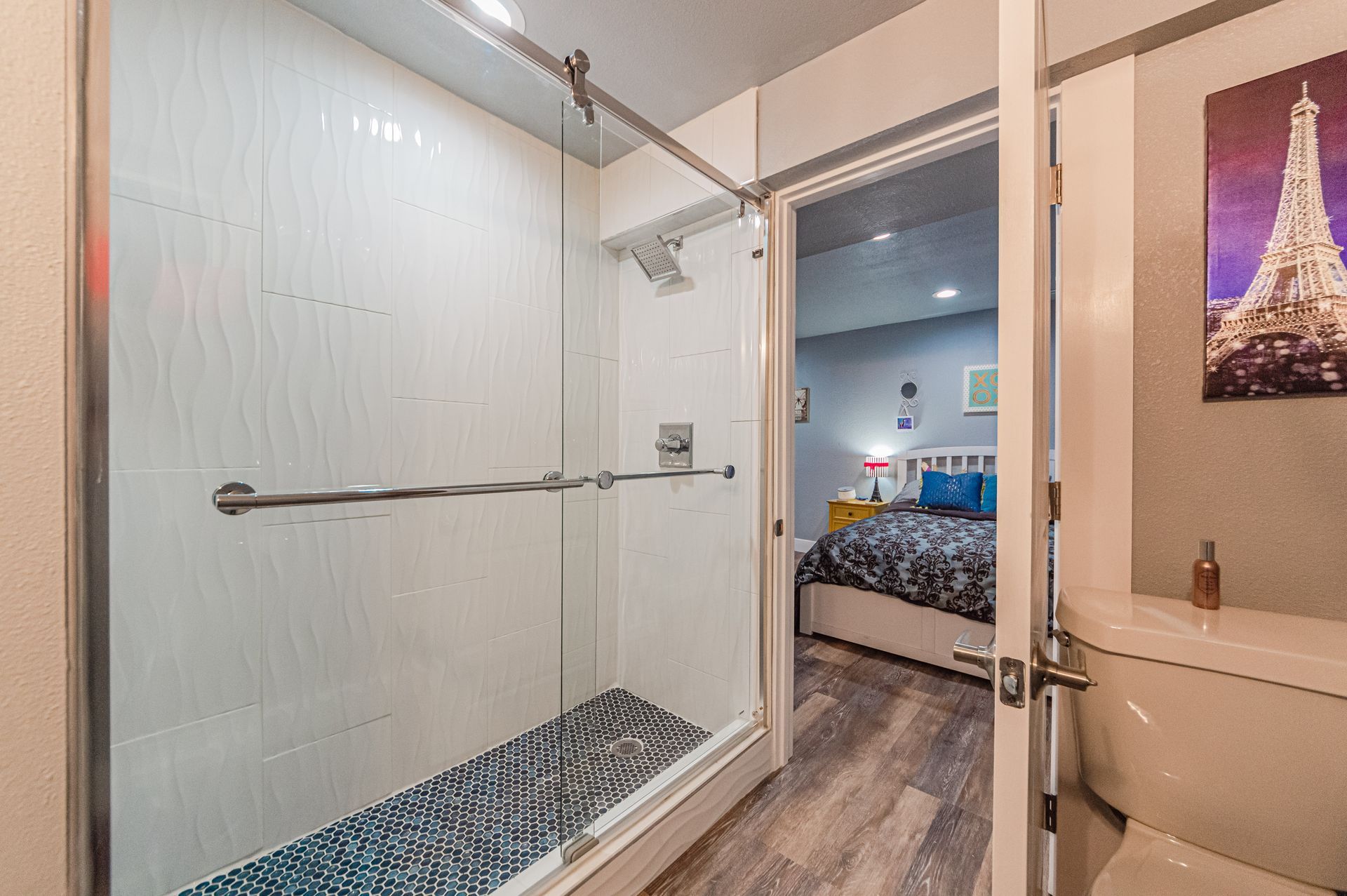 A walk-in shower with glass sliding doors and pebble floor tile, viewed from a bathroom into an adjacent bedroom.