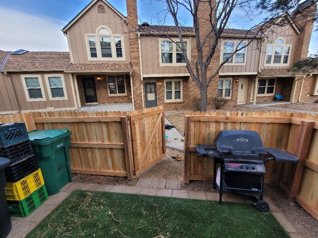 A green trash bin and a black grill sit on a patio in front of a tan brick townhouse with a wooden privacy fence.