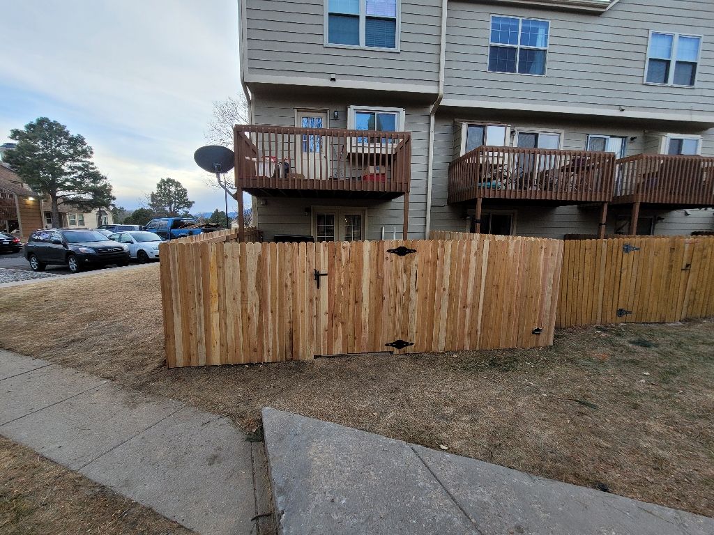 A wooden fence encloses a patio area in front of multi-story townhomes with wooden balconies on an overcast day.