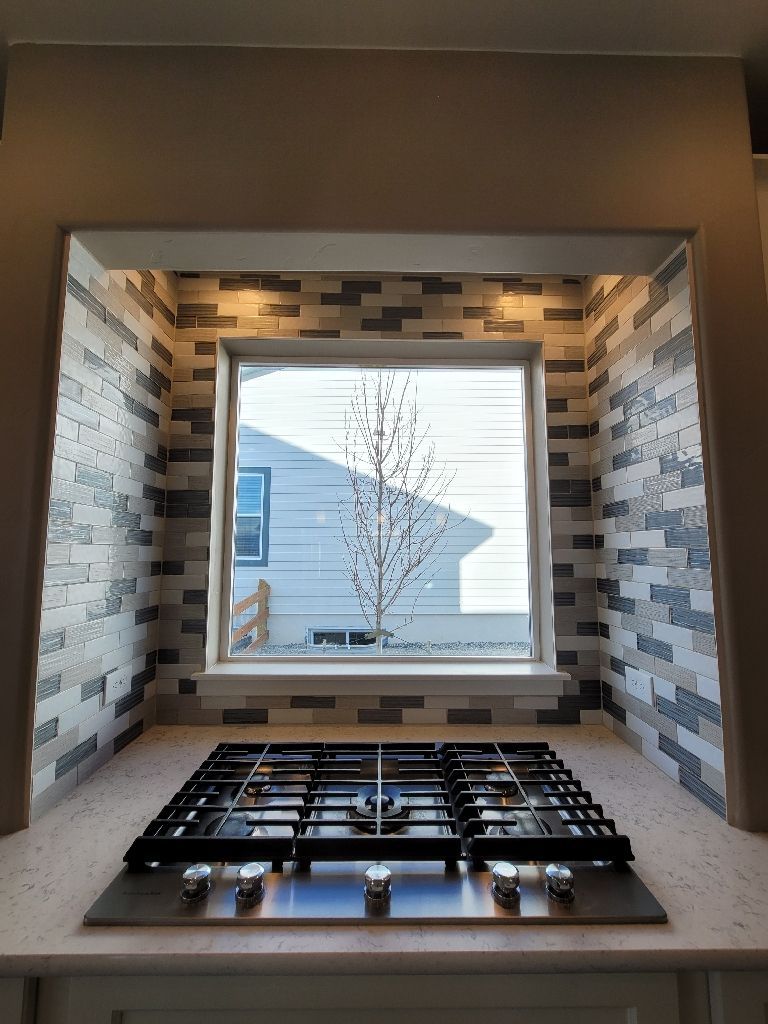 A kitchen stove top sits against a tiled wall under a recessed window overlooking a neighborhood.