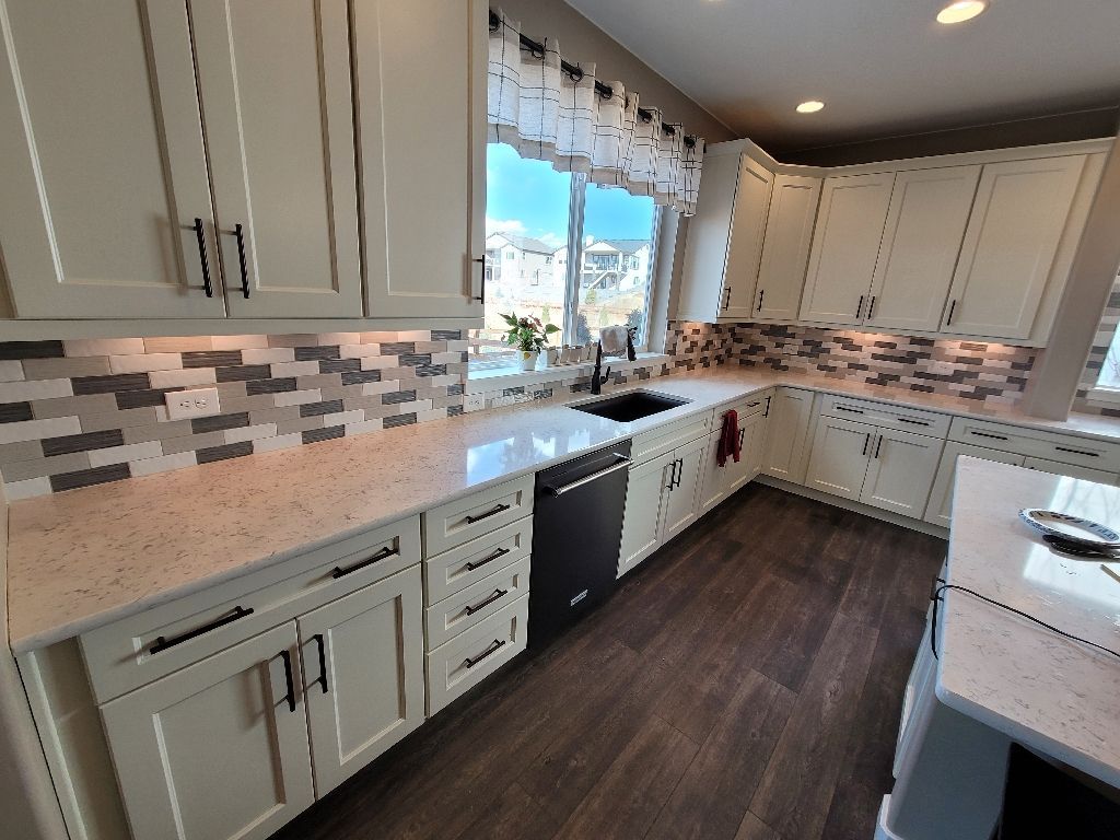 A bright kitchen featuring cream-colored cabinets, speckled countertops, a patterned tile backsplash, and dark wood floors.