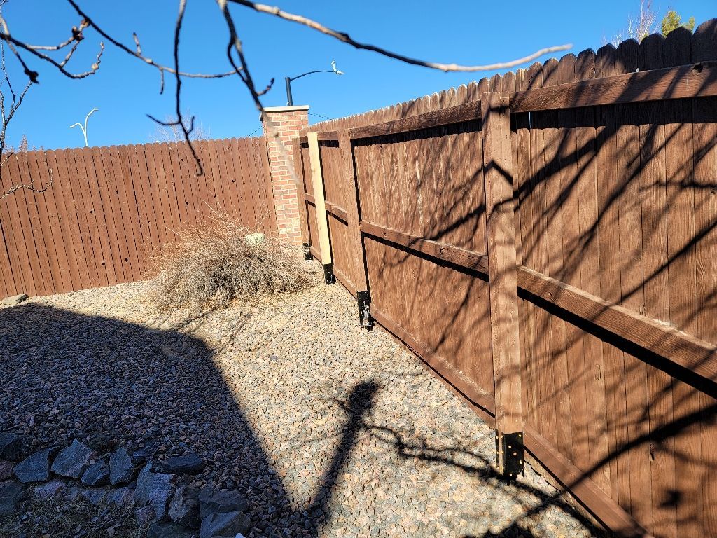 A fenced backyard area with a brick pillar, brown wooden privacy fence, and gravel ground under a clear blue sky.