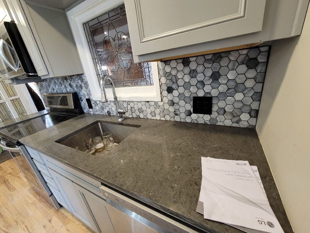 A gray kitchen with a dark stone countertop, a stainless steel sink, and a multi-toned blue and gray hexagon tile backsplash.