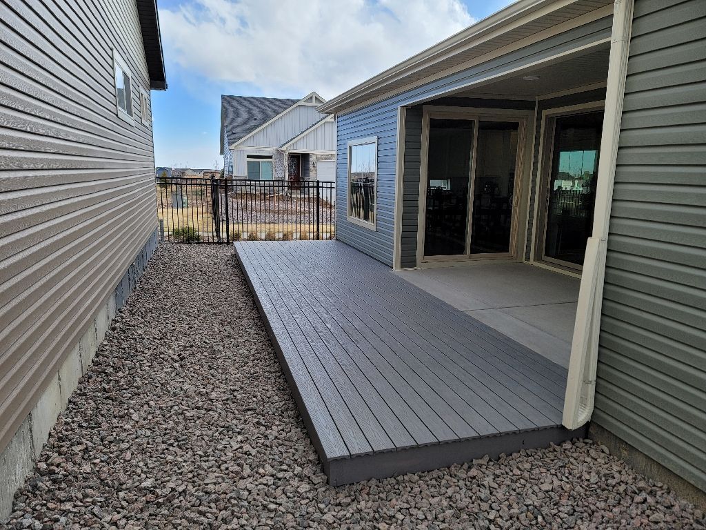 A grey wooden deck extends from a sliding glass door along the side of a house with gravel landscaping.