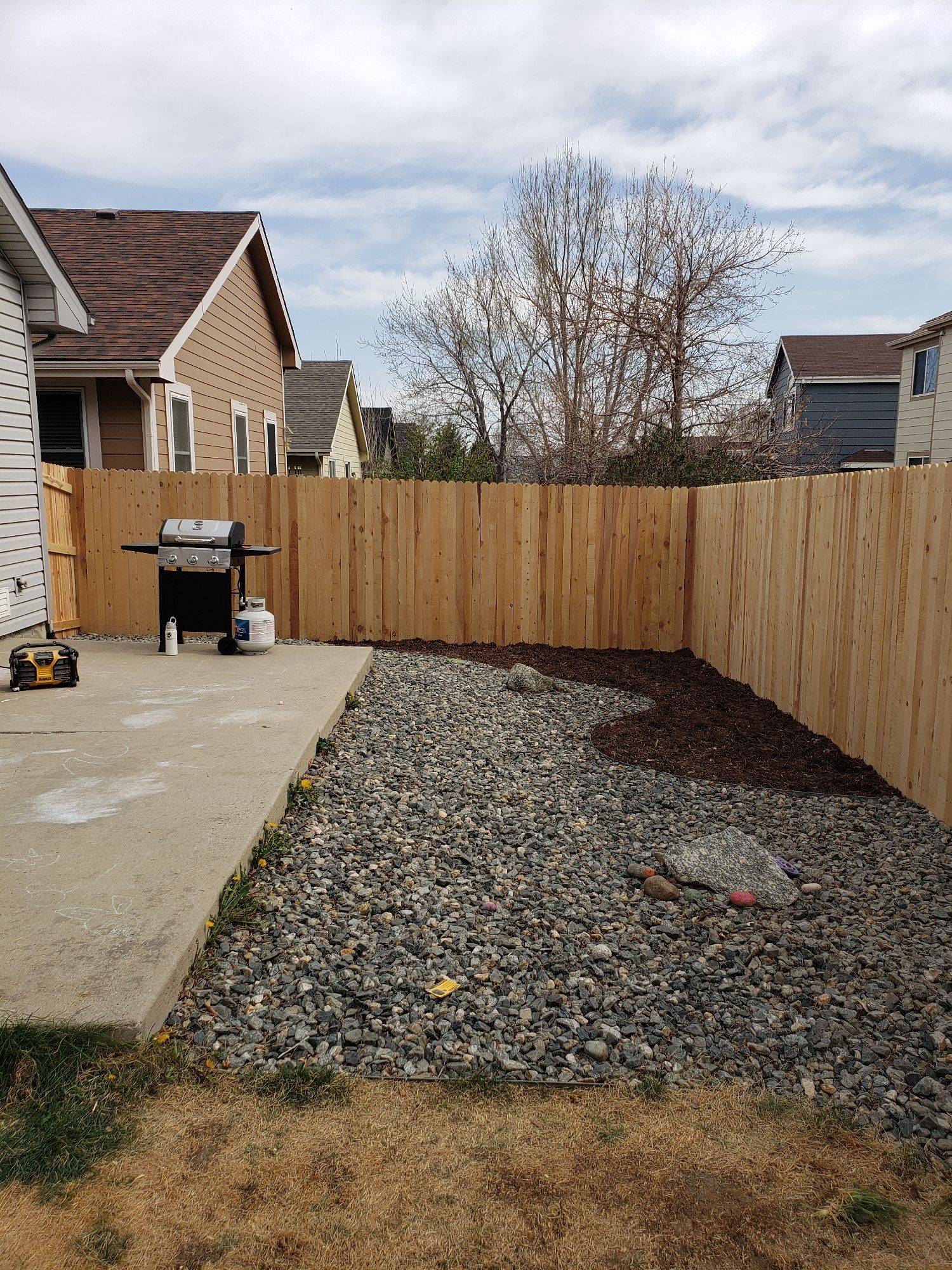 A residential backyard featuring a concrete patio, a gravel area with some mulch, and a new wooden privacy fence.