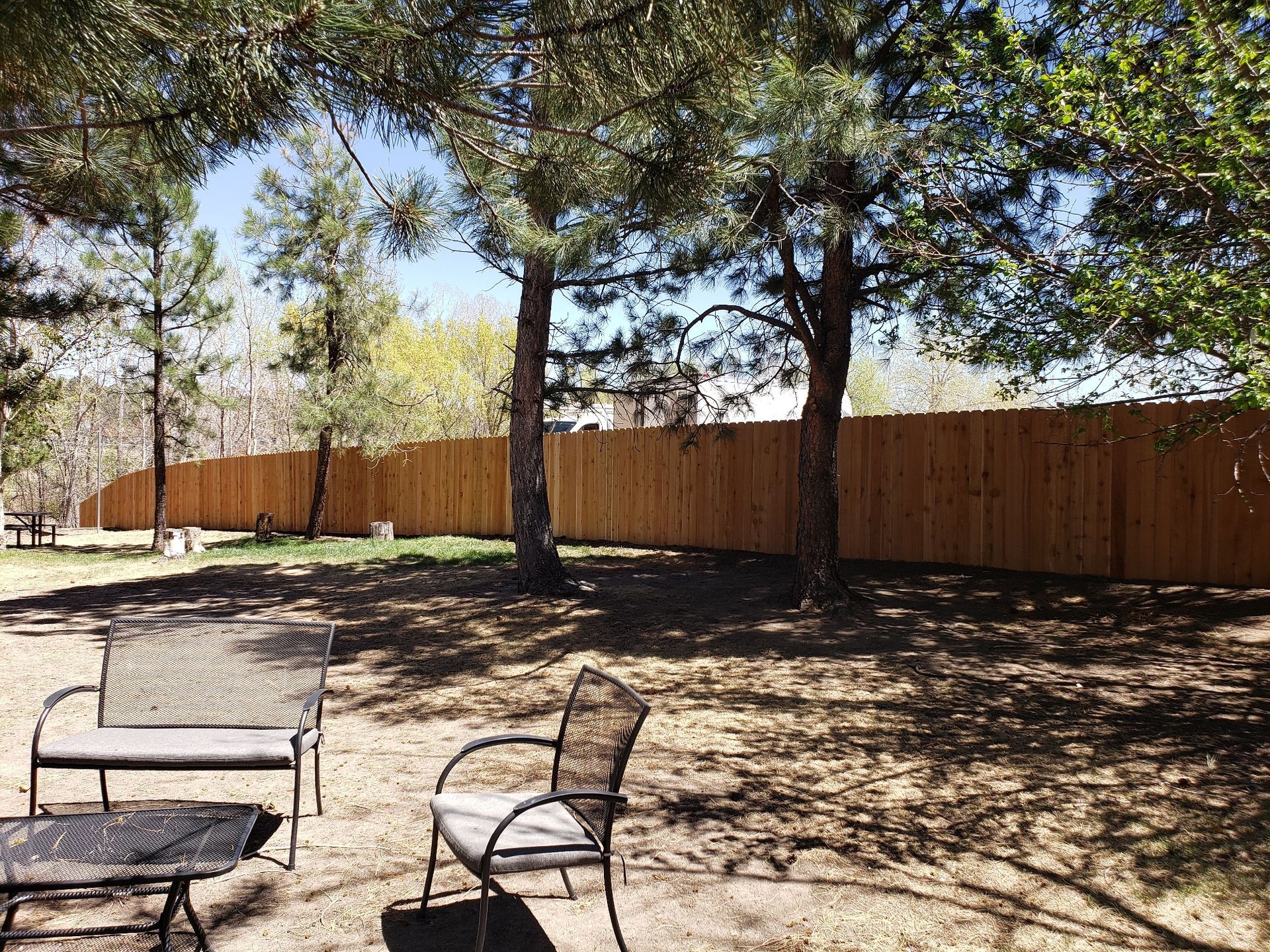 A wooden fence sits at the edge of a yard shaded by trees, with metal patio furniture in the foreground.