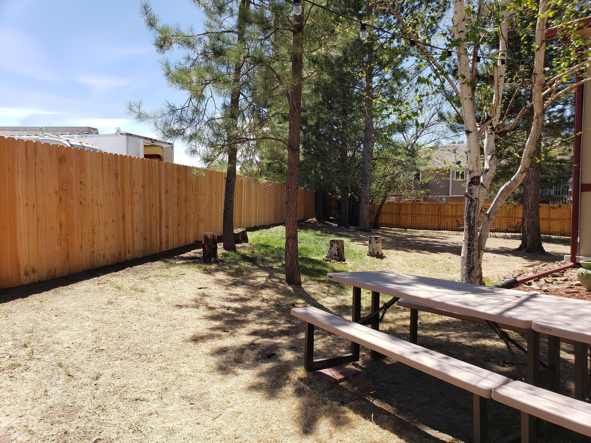 A wooden picnic table sits on a dry, grassy yard enclosed by a tall, light-brown wood fence and several mature trees.