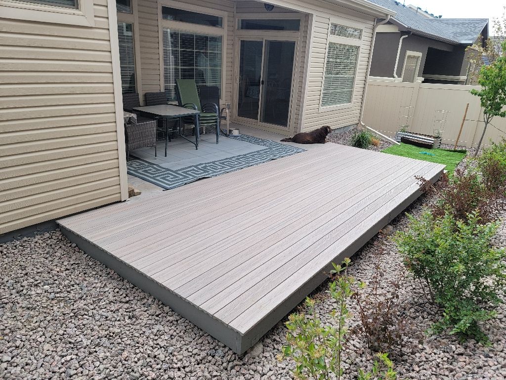 A rectangular gray deck extends from a tan house, featuring a patio set on a patterned rug and a dog resting nearby.