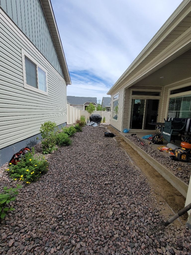 A narrow side yard between two residential houses, featuring a gravel ground cover and a concrete patio on the right.