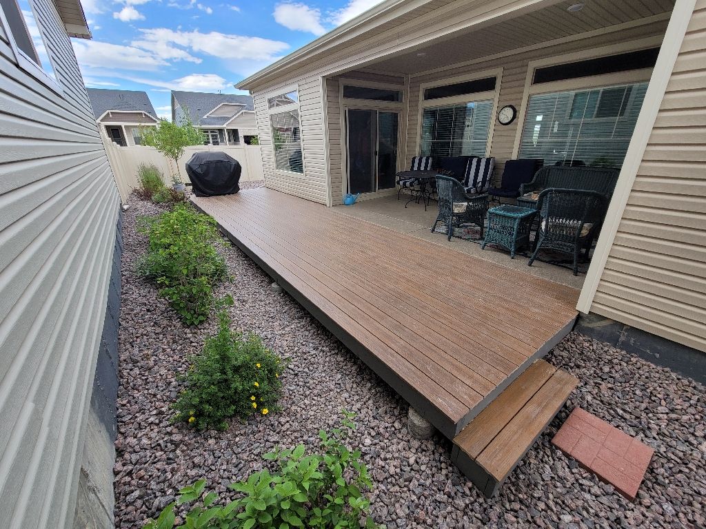 A wooden deck beside a house with beige siding, featuring outdoor furniture, a covered grill, and a gravel yard.