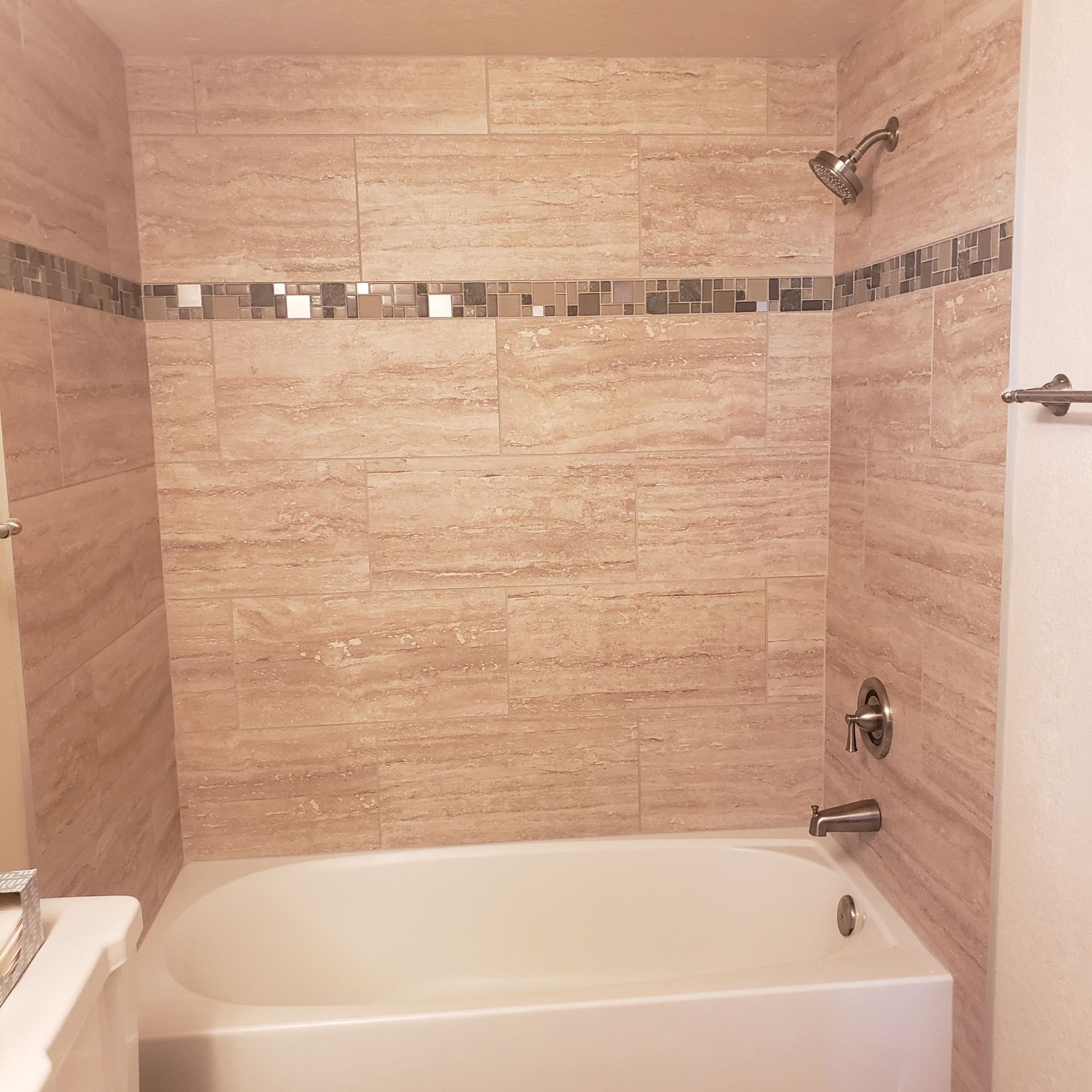 A tan-tiled bathtub and shower surround with a horizontal decorative glass mosaic accent tile strip.