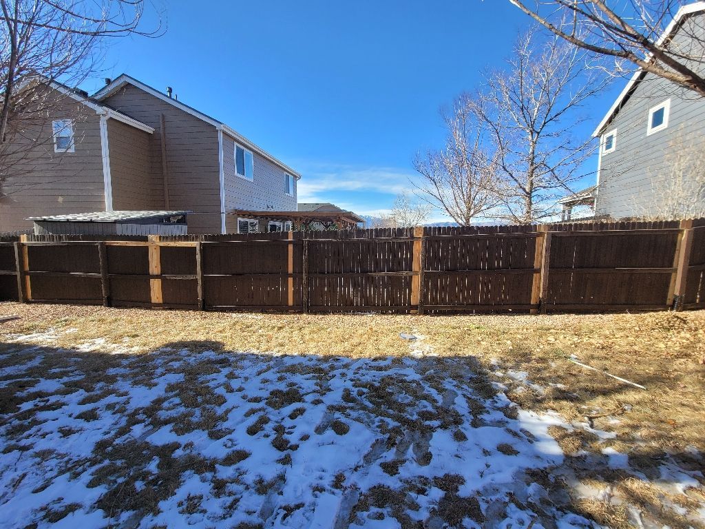 A brown wooden fence runs across a snow-dusted backyard, framed by two neighboring houses against a clear blue sky.