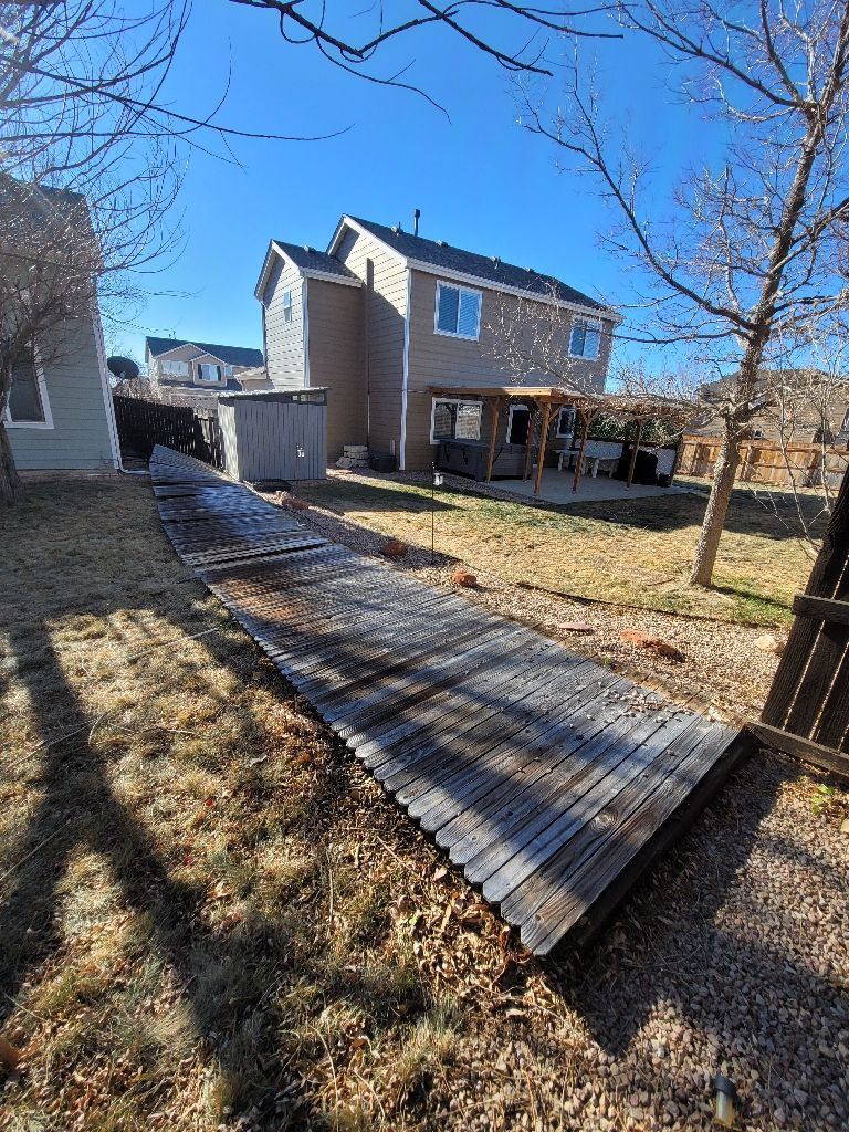 A fallen wooden fence section lies flat on the ground in a residential backyard next to a two-story beige house.